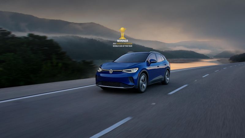 A VW ID.4 EV in Metallic Dusk Blue drives on a two-lane highway with mountains in the background. Above the car sits a small version of the 2021 World Car Awards logo and type treatment that reads “Winner” of “World Car of the Year”