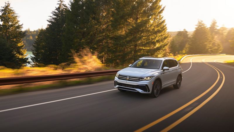 A Tiguan in Oryx White rounding a corner on a two-lane road with trees in the background, as seen from the front driver side.
