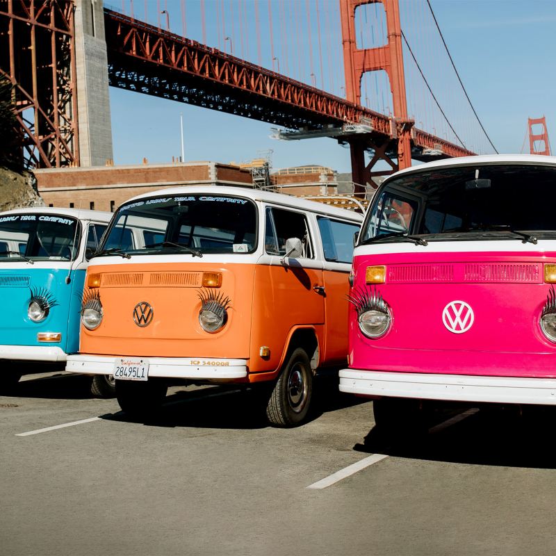 Four vintage Volkswagen buses sit in front of a bridge