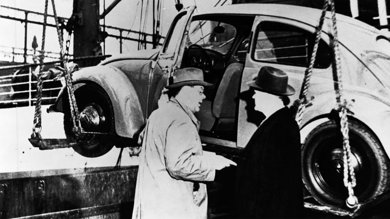 Two men stand in front of an early model of the Volkswagen Beetle.