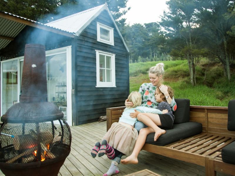 A woman and her children sit in front of an outdoor fireplace outside a tiny home.