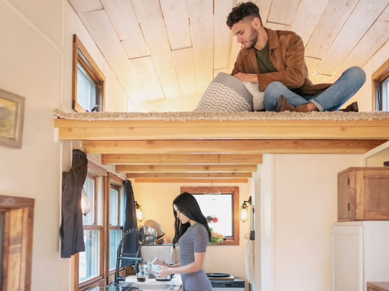 A man sits in a loft while a woman stands on the first floor below, inside a tiny house.