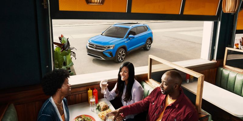 Tres personas sentadas en un gabinete de una cafetería; se ve la parte delantera del lado del conductor de un Taos en color Cornflower Blue por la ventana.