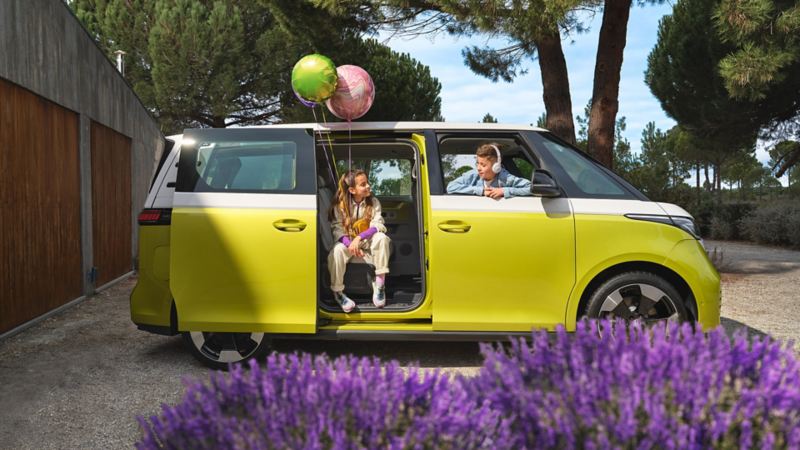 A profile shot of an ID. Buzz in Candy White and Lime Yellow parked on a driveway. A girl holding two balloons sits facing out from the open  side-pocket door. She is looking at a boy wearing headphones who is looking back at her through the open passenger window.