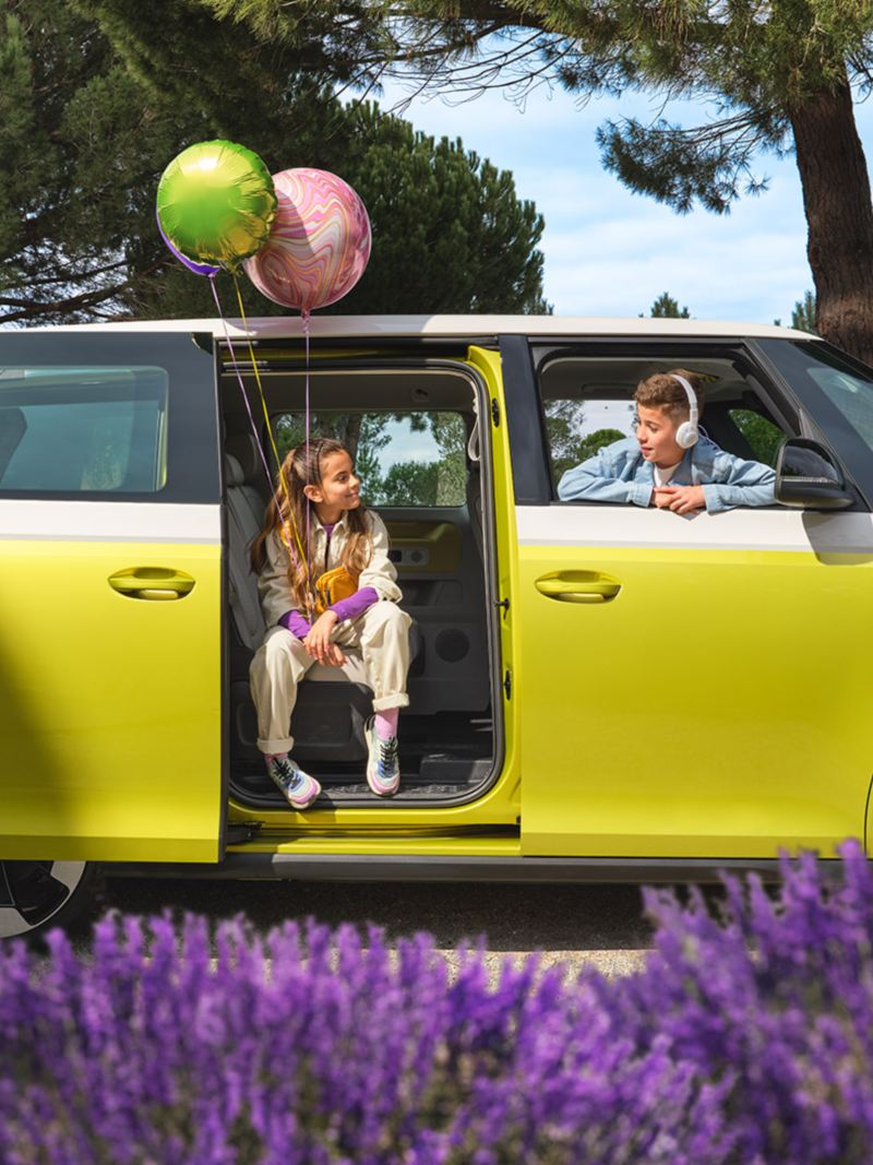 A profile shot of an ID. Buzz in Candy White and Lime Yellow parked on a driveway. A girl holding two balloons sits facing out from the open  side-pocket door. She is looking at a boy wearing headphones who is looking back at her through the open passenger window.