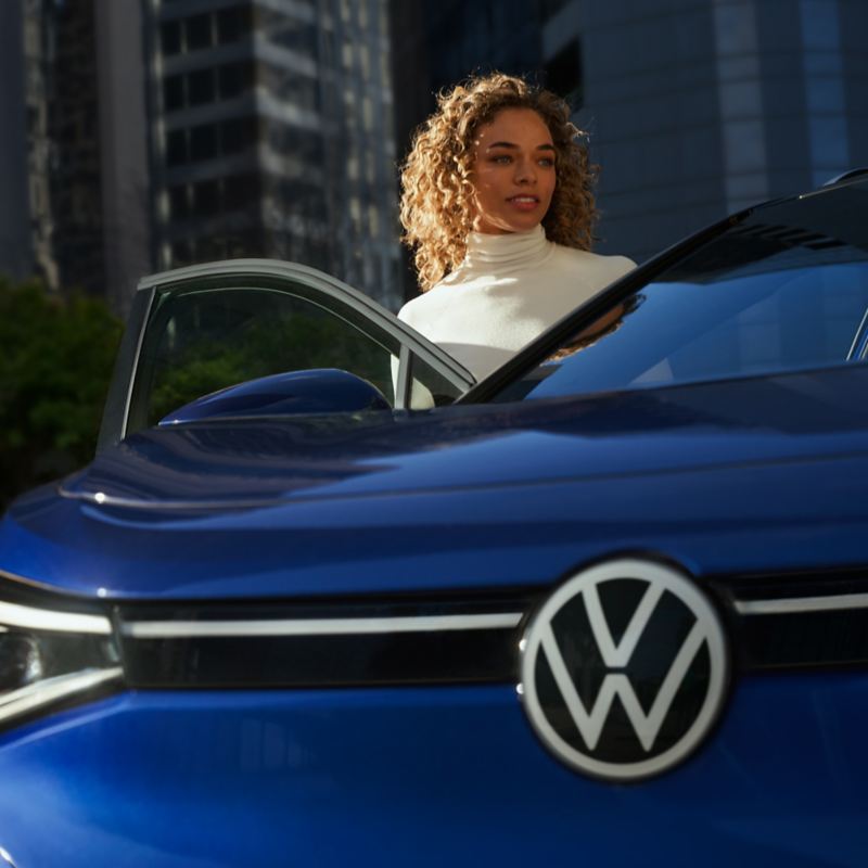 From a low angle, up past the VW grille emblem and over the hood, a woman stands inside the open passenger door of an ID.4 in Dusk Blue Metallic.