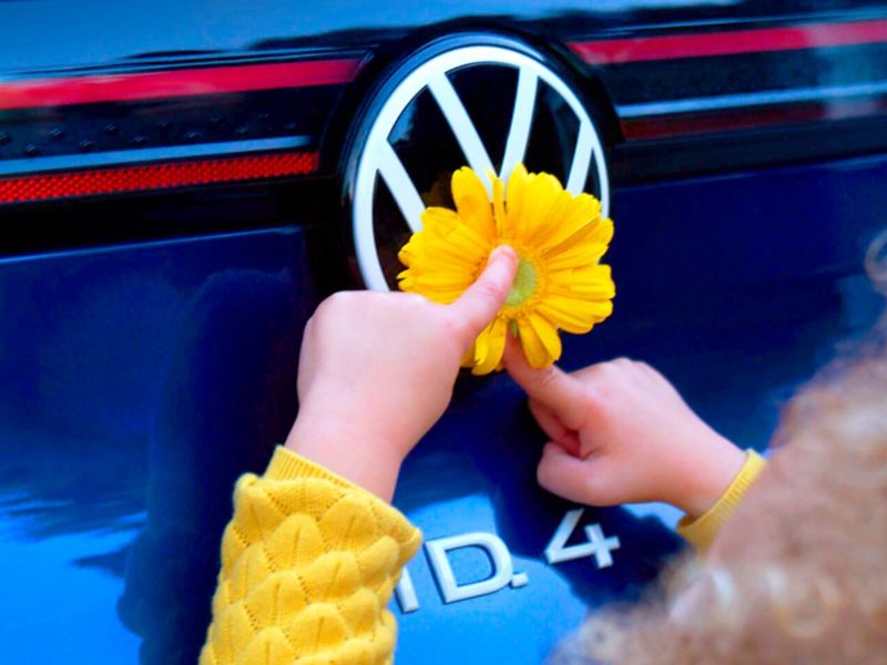 Un niño pequeño sostiene una flor sobre el logotipo de VW de un ID.4 en color Dusk Blue Metallic.