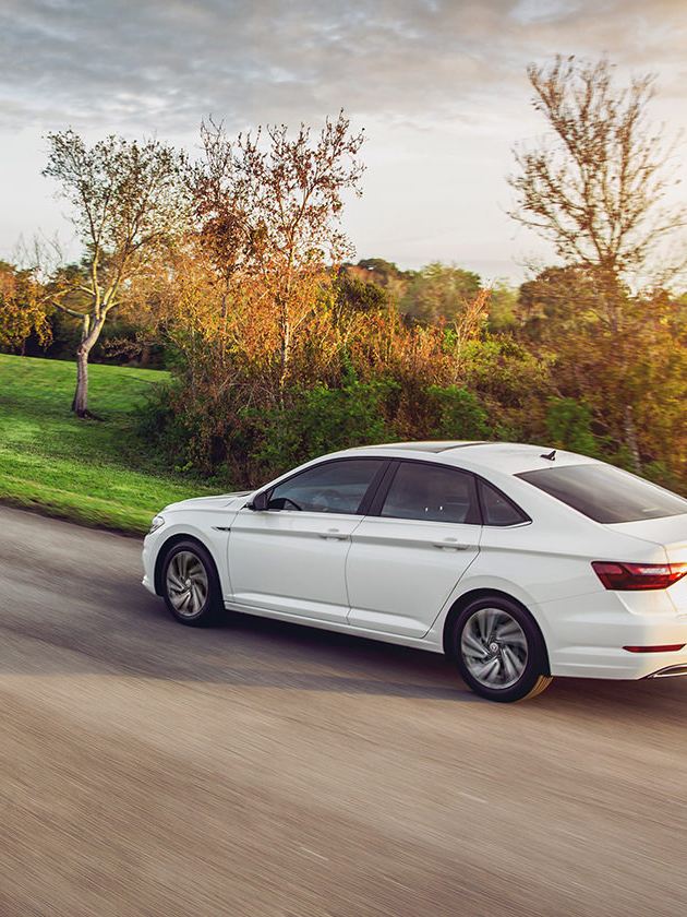 Jetta shown in Pure White driving on a road as seen from the rear with grassy hills in the distance.