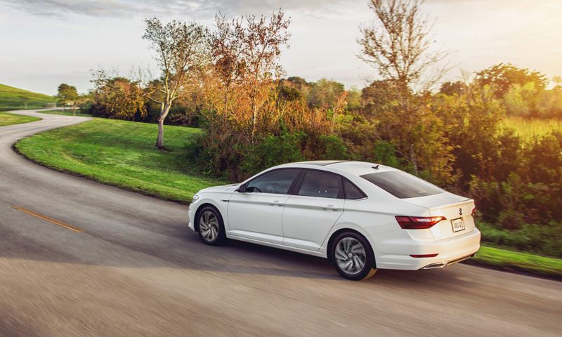 Jetta shown in Pure White driving on a road as seen from the rear with grassy hills in the distance.