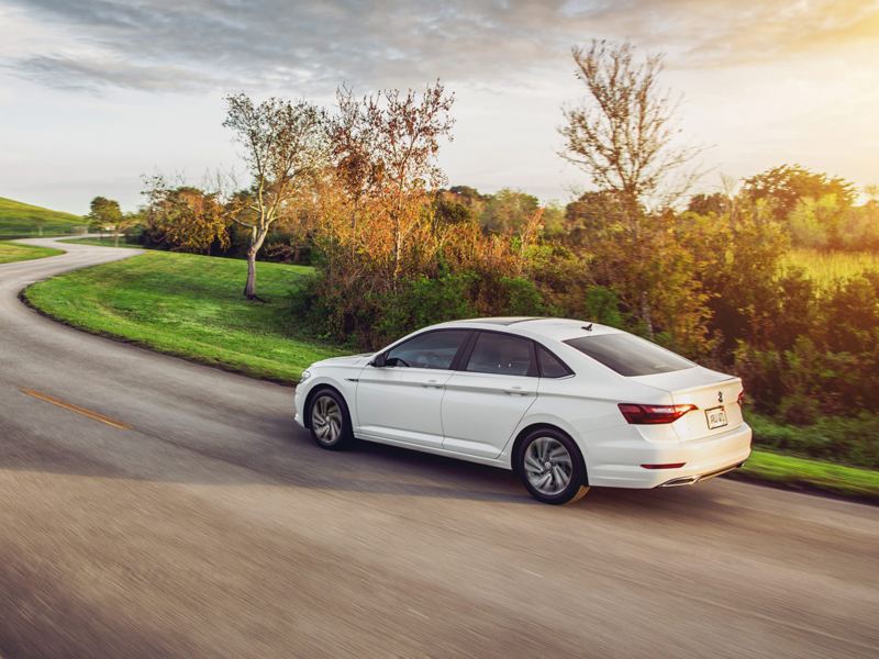 Jetta shown in Pure White driving on a road as seen from the rear with grassy hills in the distance.