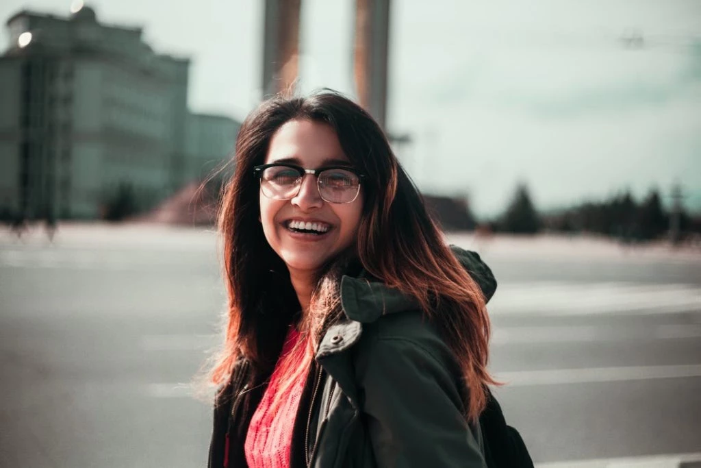 Woman standing in city with long reddish hair smiling, wearing green jacket and a red shirt.