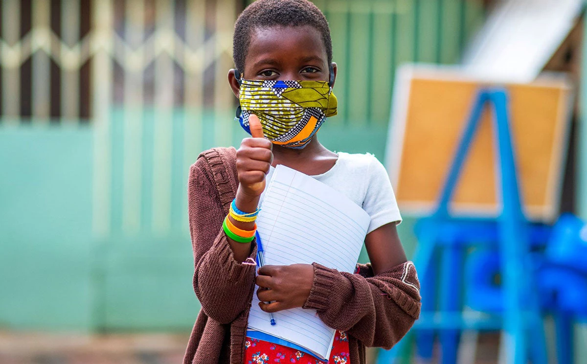 Una niña africana usa una mascarilla hecha en casa como protección y da su aprobación levantando el pulgar derecho, con la escuela en segundo plano (crédito de la fotografía: Yaw Niel)