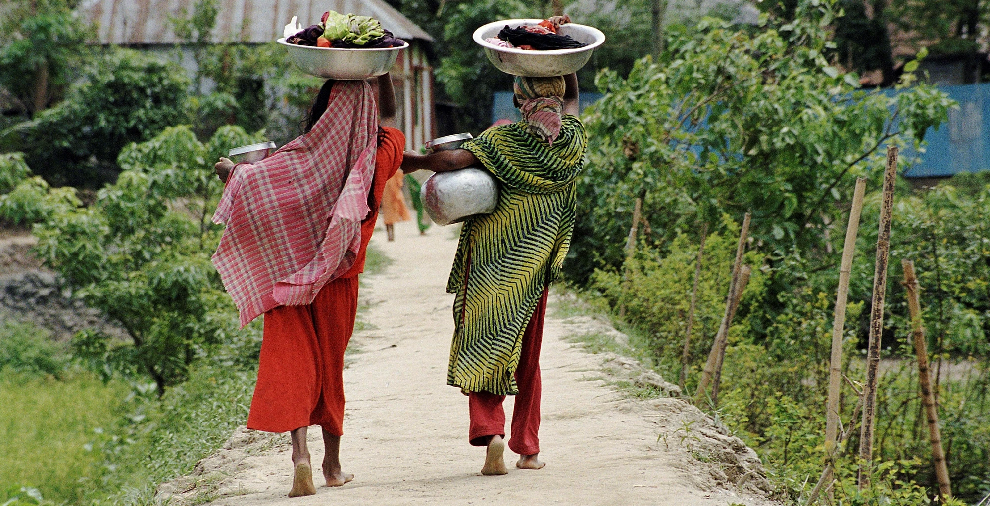 Two women carry clothing, produce, and water jugs in Bangladesh.
