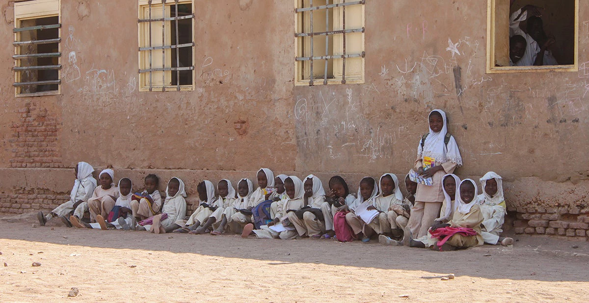 Children seated on the floor outside, leaning against the wall of their school