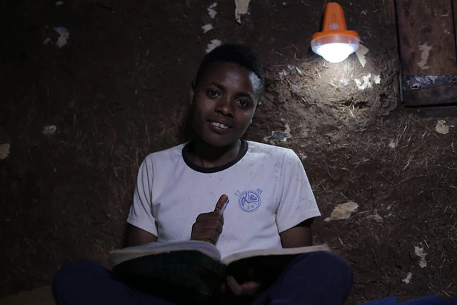 A child holds a notebook and pen under a solar-power light in a dark room