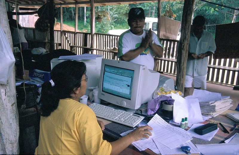 A woman in a yellow shirt sits behind an old computer, while a man stands across from her waiting