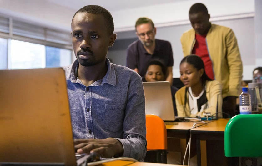 Photo of an adult African man in front of a laptop, with a group of students behind him on their computers.