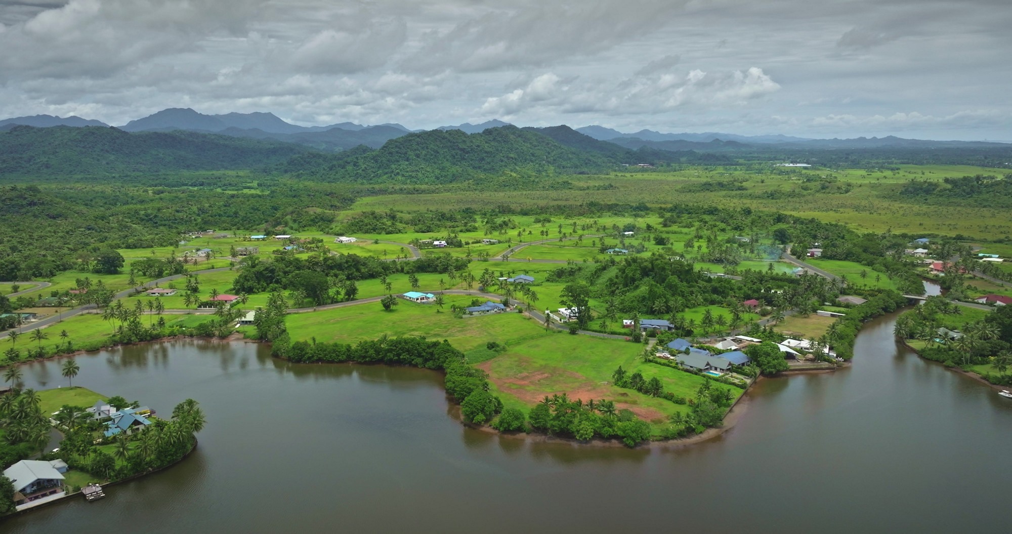 Drone capturing breathtaking scenery of Savusavu town featuring lush green landscape, meandering river, residential houses, and rolling hills in Fiji, showcasing tropical beauty and tranquility