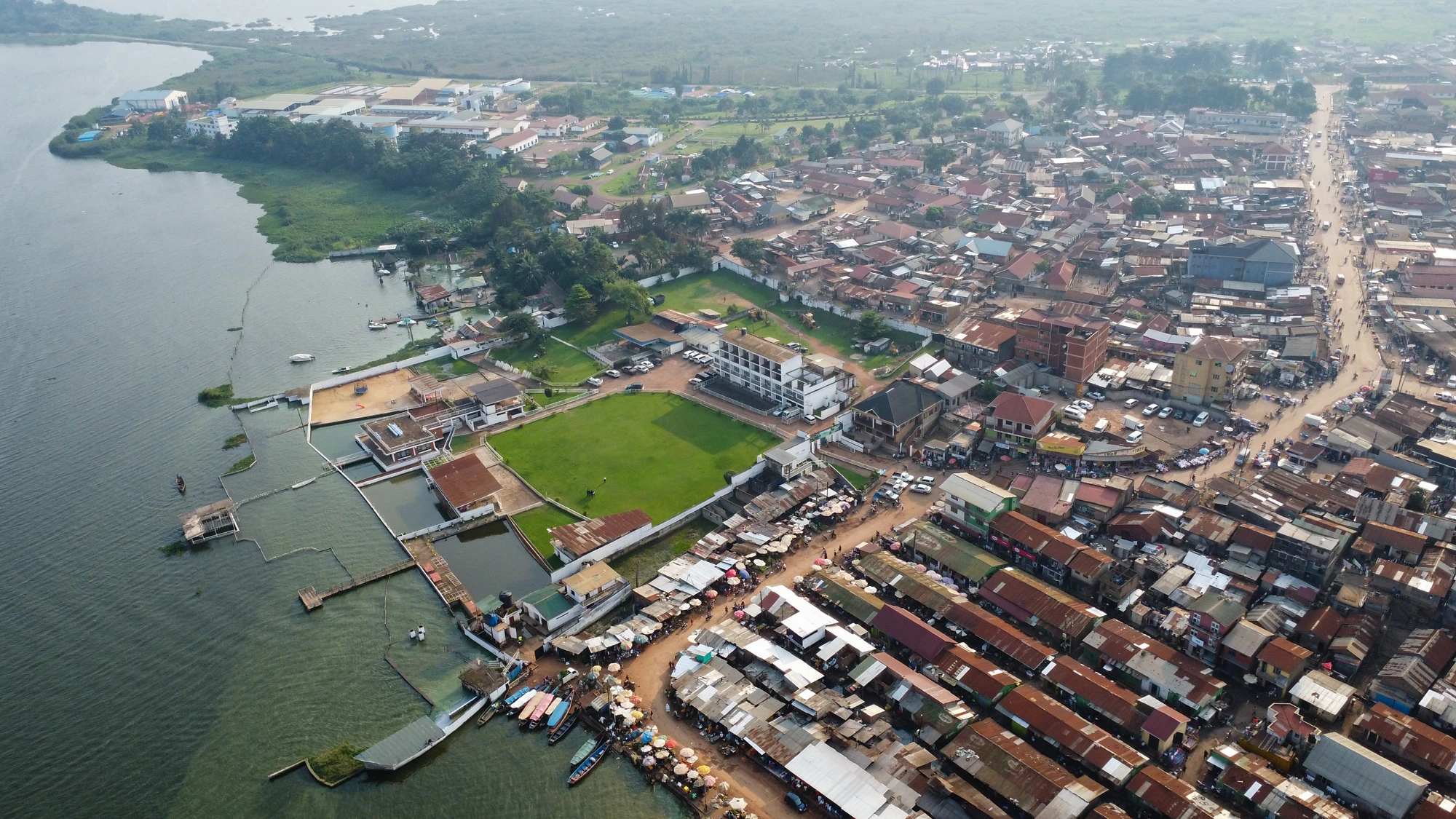 This aerial photograph showcases Jinja, Uganda, with its dense residential buildings, Lake Victoria waterfront, and a bustling market area alongside the main road. The image also features green spaces and various infrastructure elements.
