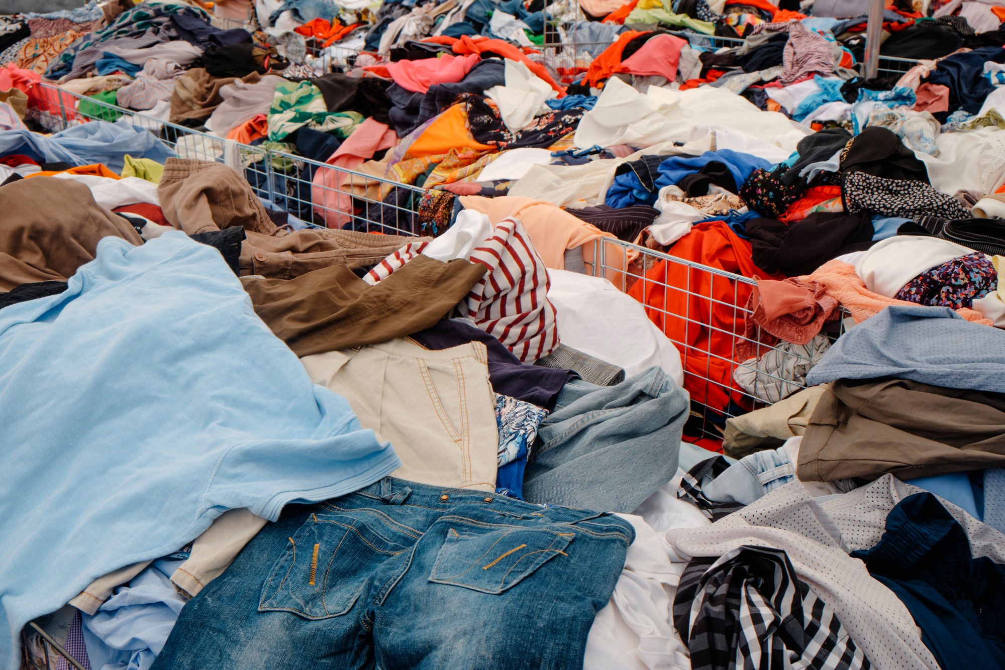 piles of clothes in assorted colors and fabrics are stacked inside metal bins at an outdoor market stall full of mixed garments on display