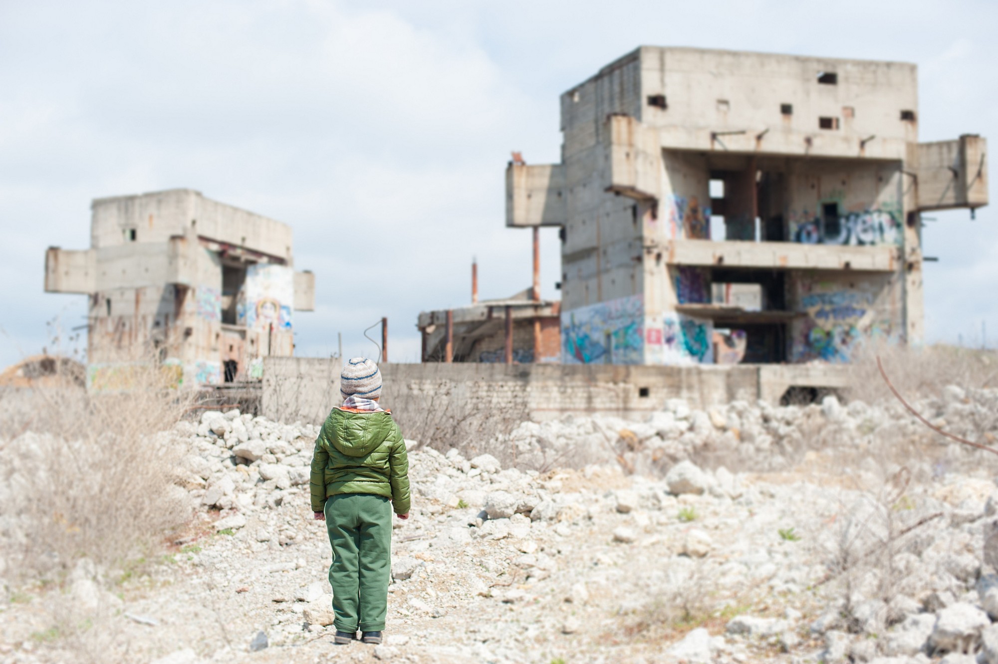 little boy in jacket stands against ruins of building as result of war conflict