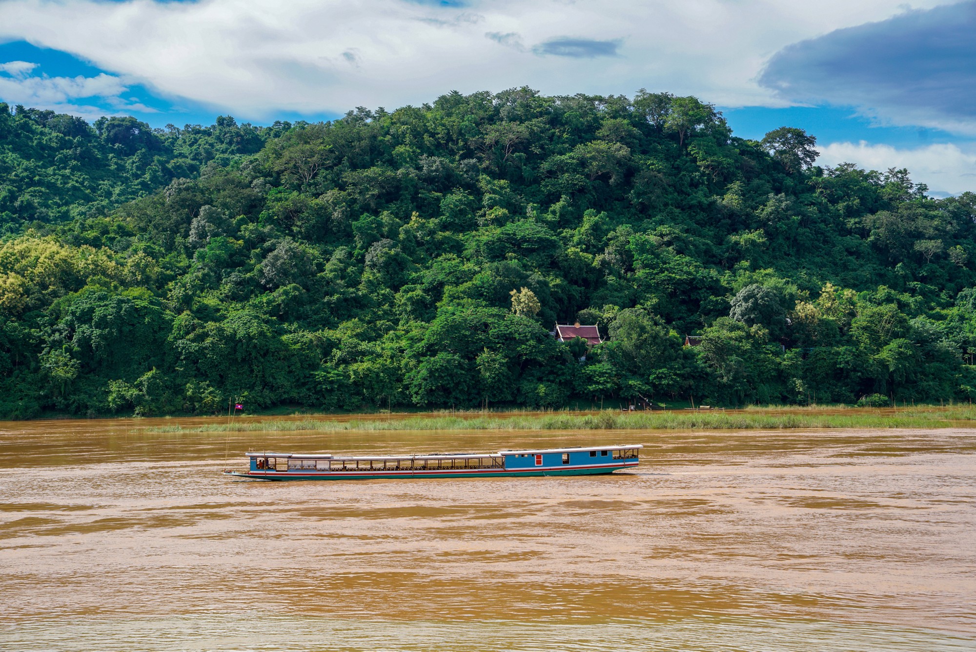 A traditional Laos slow boat cruising a muddy brown lower basin Mekong River after high rainfall. Tropical jungle covers the mountainous background of the UNESCO World Heritage Site of Luang Prabang