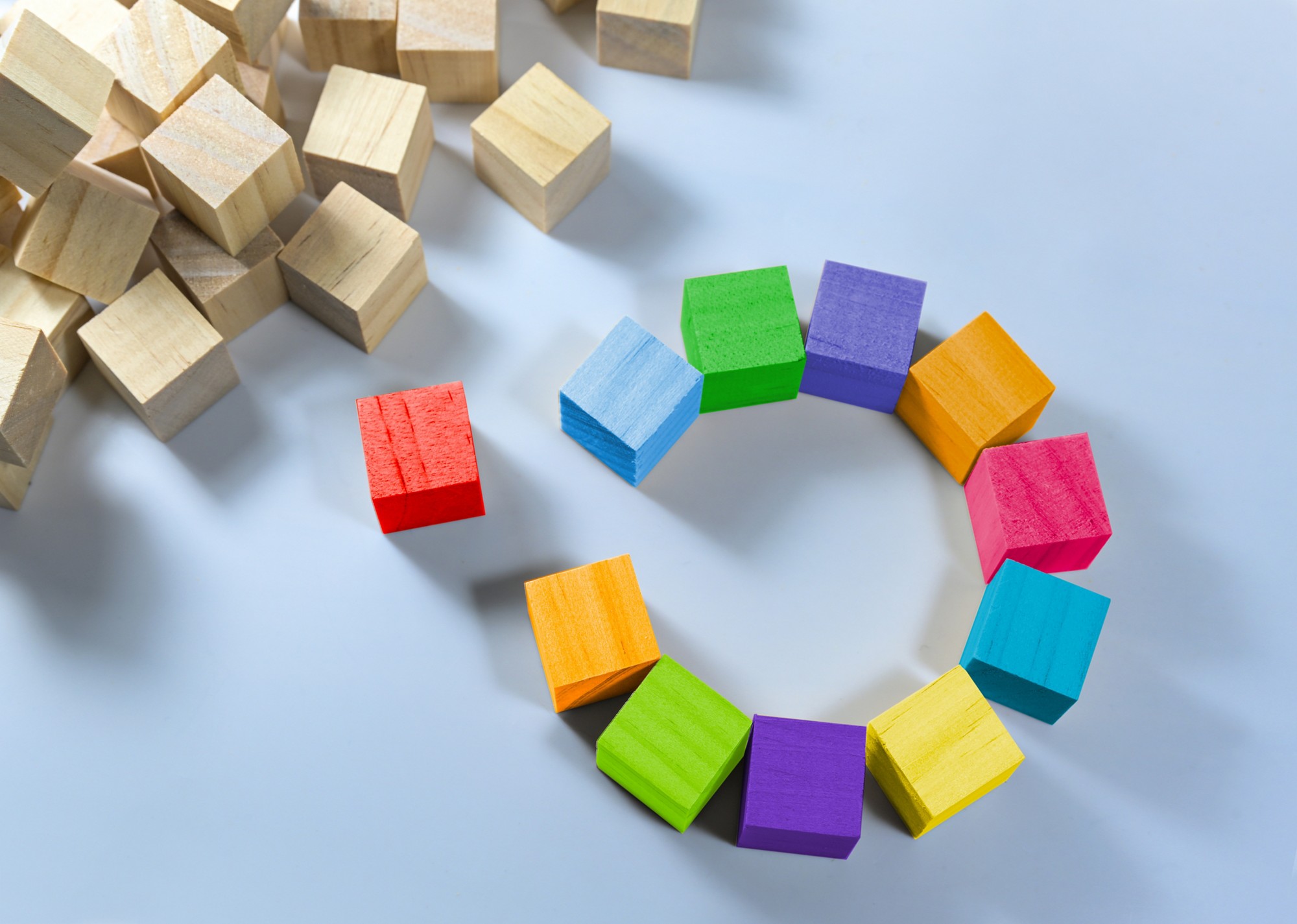 Cubes in different colors together form a circle with a gap, one is red and stands separate, heap of natural wooden cubes in the corner, light blue background, copy space, selected focus