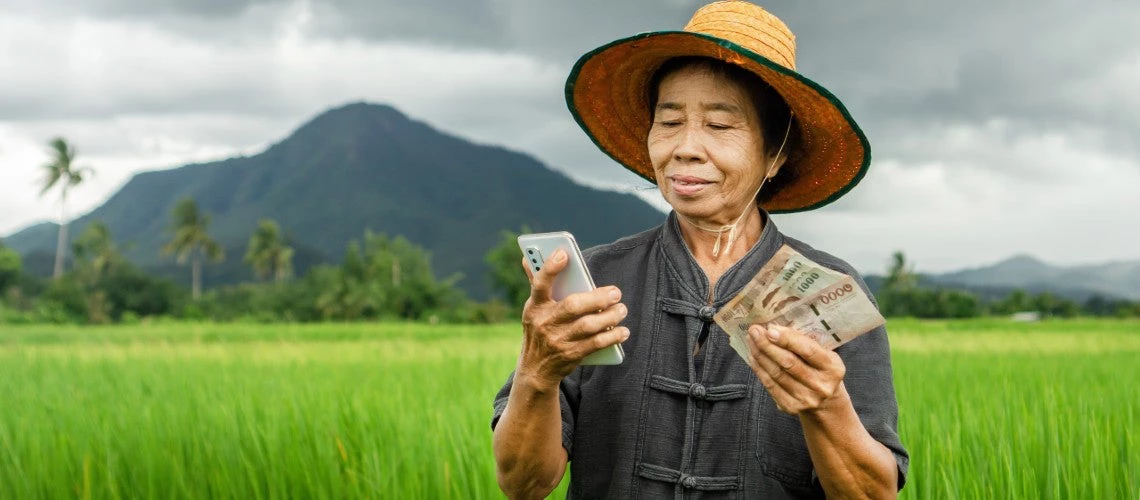 Smiling Asian woman farmer holds Thai banknotes and a mobile phone, illustrating the benefits of cash subsidy