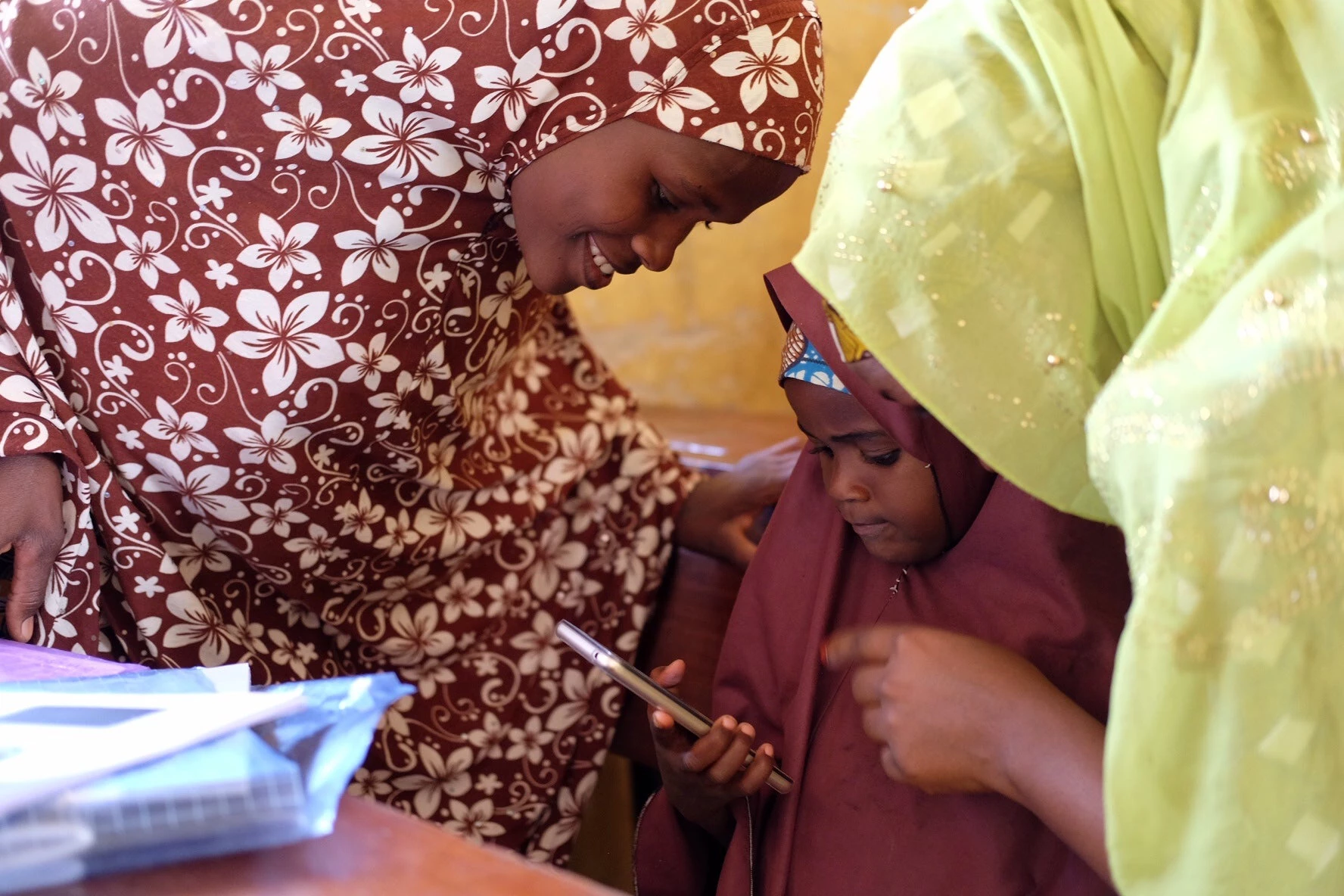 An African female teacher proudly smiles at her young female student who uses a smartphone to learn in the classroom