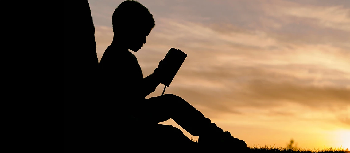 A silhouette of child reading under a tree during sunset. | © Aaron Burden / Unsplash