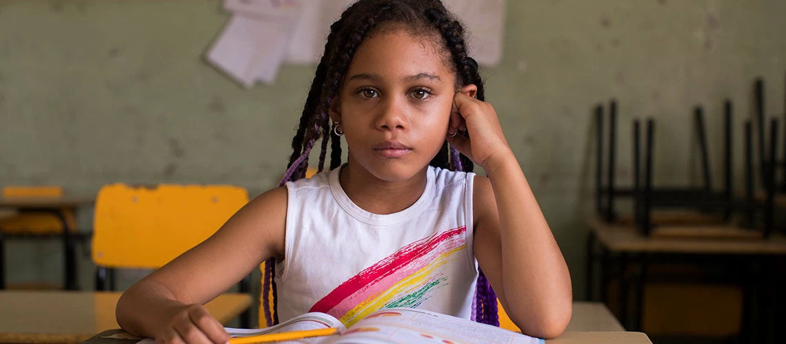 Niña afrodescendiente en una aula de clase con lapiz en la mano y un libro sobre el pupitre 