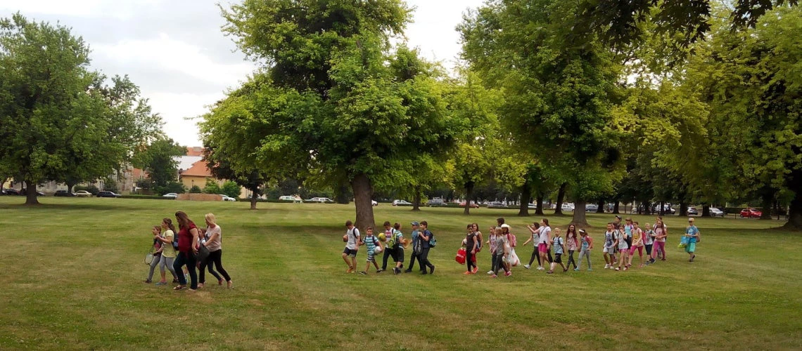 Group of children in Croatia accompanied by teachers walking on grass through a park towards their playground.