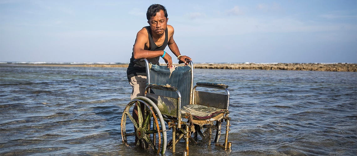 A man standing in the water with a wheelchair of his grandmother in Jenrok village, Majuro in the Marshall Islands.