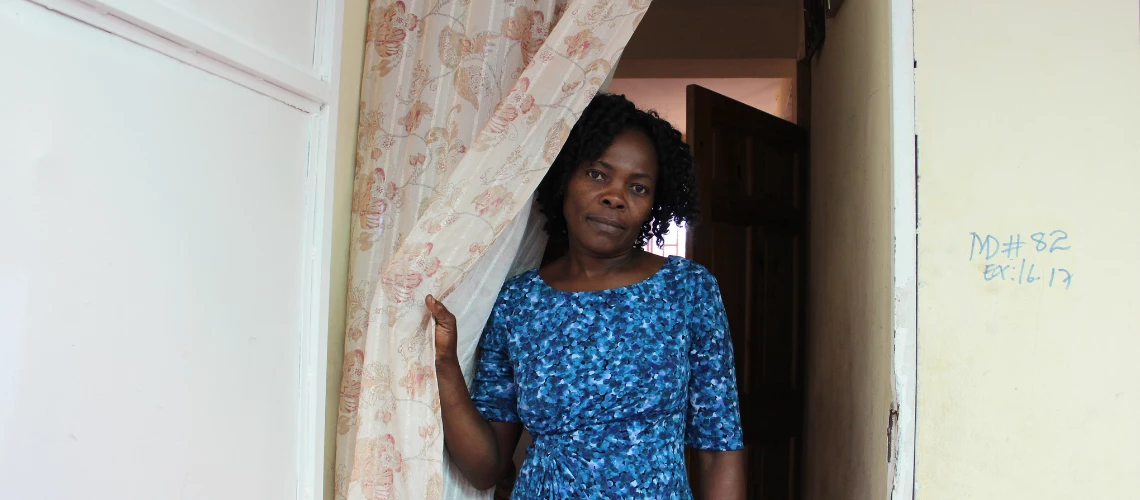 A resilient Haitian woman gazes from the doorway of her home, clutching onto a vibrant curtain.