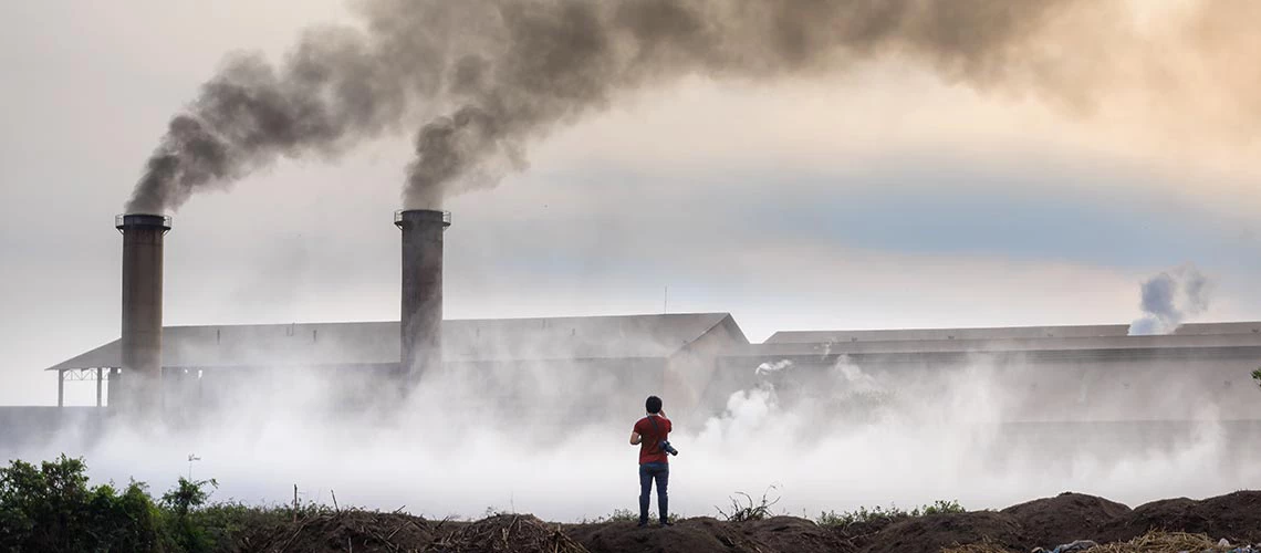 A photographer facing a factory with two big chimneys and grey smoke coming out of them. 