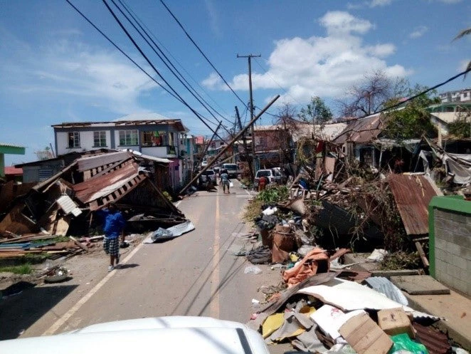 People walking in the street with destroyed houses on either side of them.