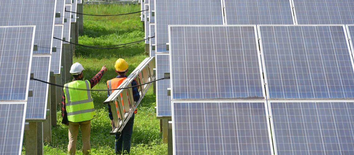 Two male electrician workers walking in between long rows of photovoltaic solar panels and talking about installation of new solar panels. Photo credit: Shutterstock
