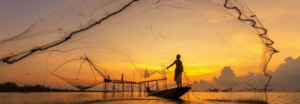 A young fisherman in Asia casts a net at dawn as the sun rises behind him. 