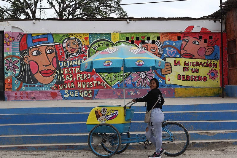 Young street vendor with a bicycle in front of a mural with messages about Venezuelan migration