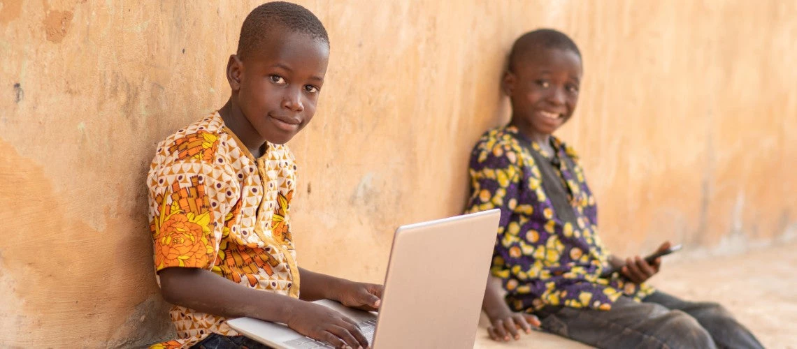 Two Mozambican children each hold a laptop and a cellphone while smiling at the camera