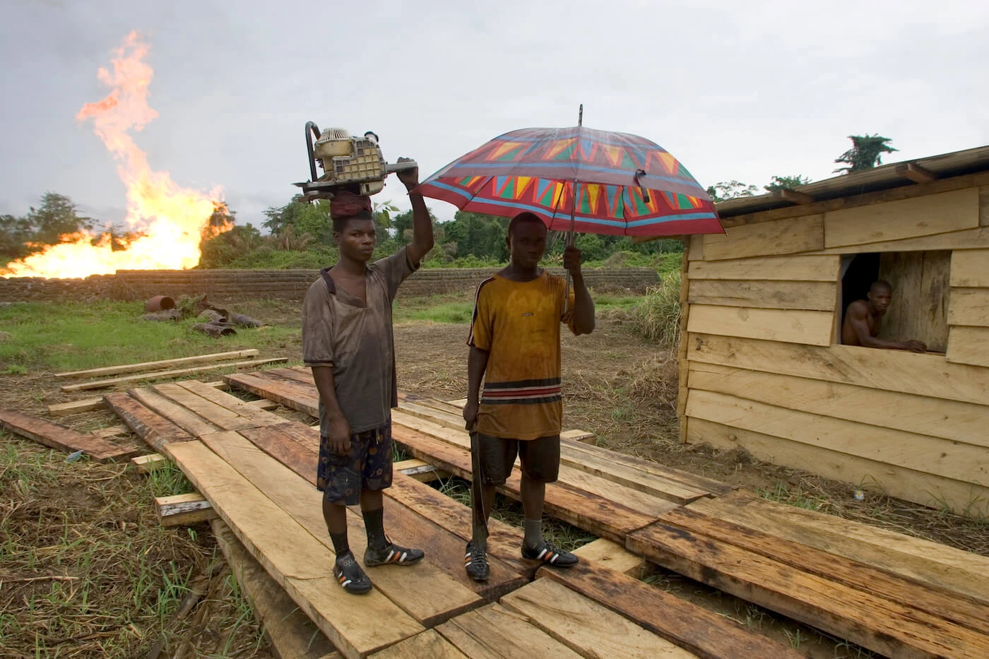 Behind three Nigerian men, a gas flaring furnace amid green fields is seen