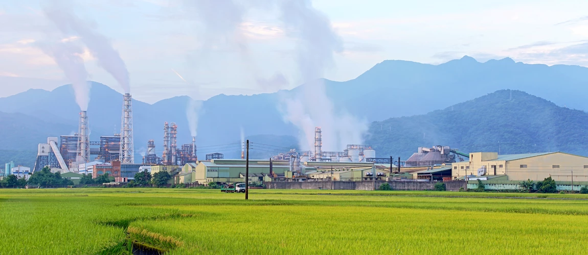 A chemical factory emitting smoke in the middle of a green rice field on a cloudy day