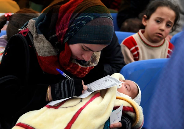 Veiled woman holding baby and filling out paperwork with another child looking at her from behind.