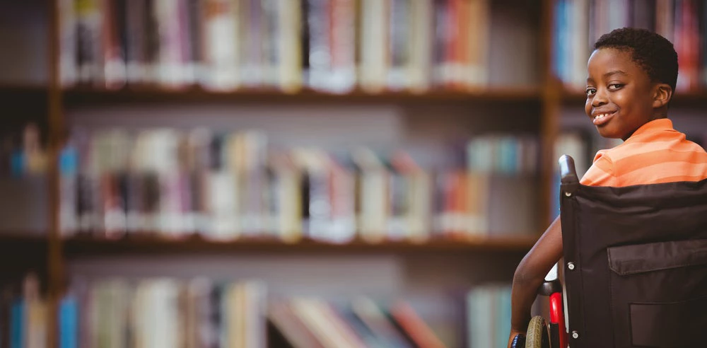 Portrait of cute boy sitting in wheelchair against library shelf by wavebreakmedia.