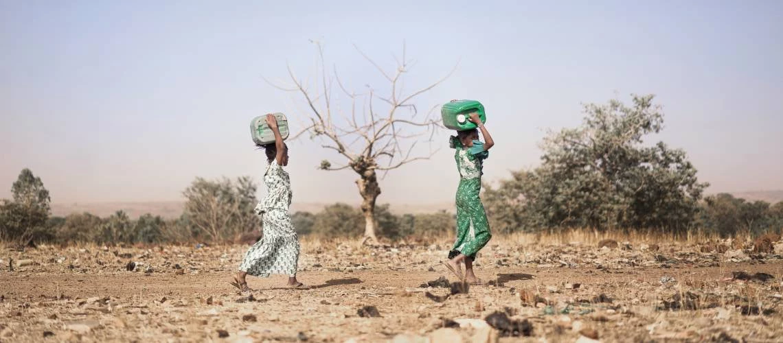 Two young girls carry containers of fresh water across dry land.