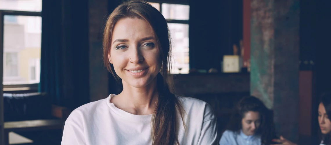 A young female office worker standing in workplace smiles while her colleagues are busy working in background