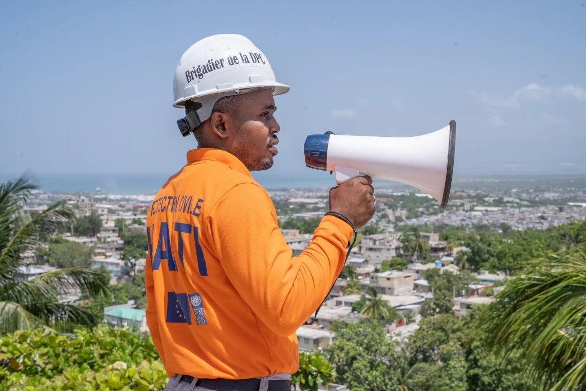 A man in an orange shirt and hard hat holding a bullhorn on a hill overlooking a city.