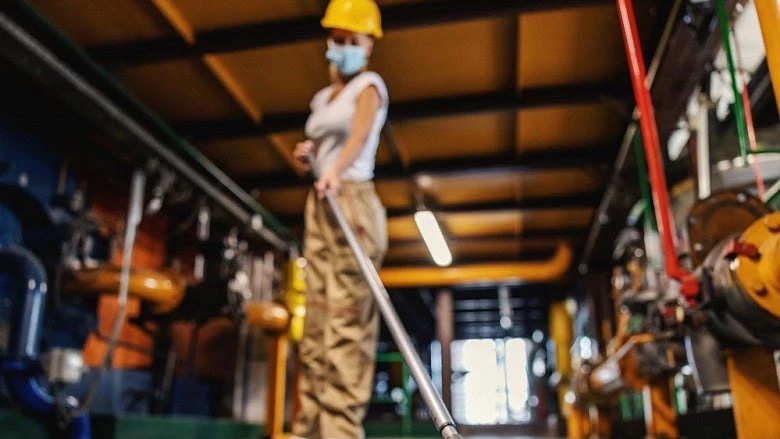 Tidy hardworking female worker in working suit with protective helmet on head and face mask brooms heating plant facility during corona outbreak.