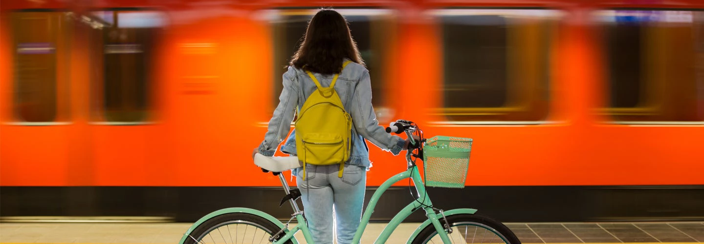 Teenager girl in jeans with yellow backpack and bike standing on metro station, waiting for train.