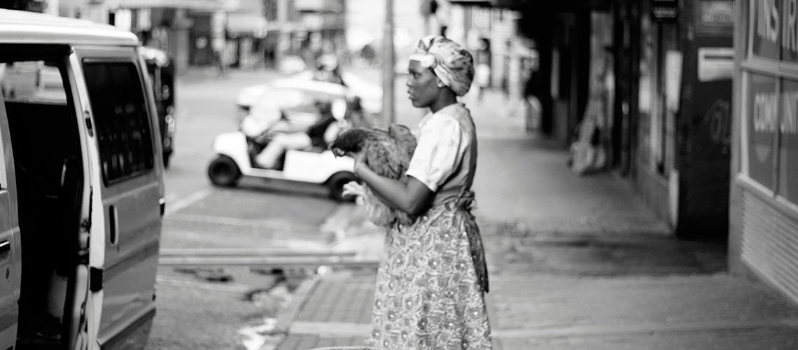A grayscale photo of woman holding a chicken standing on a sidewalk. | © Hennie Stander / Unsplash